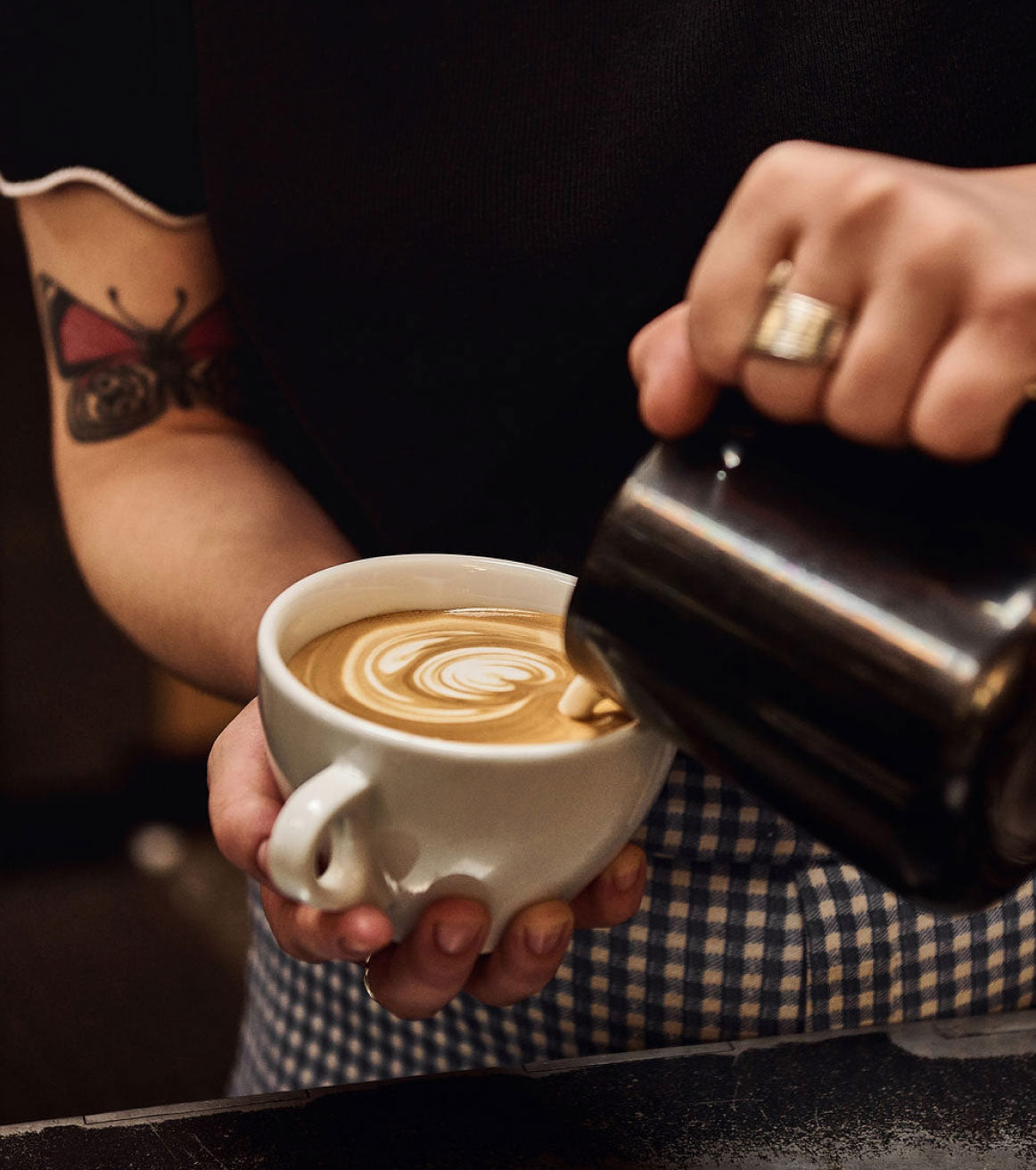 Person pouring coffee into a white cup with a tattooed arm and checkered sleeve.